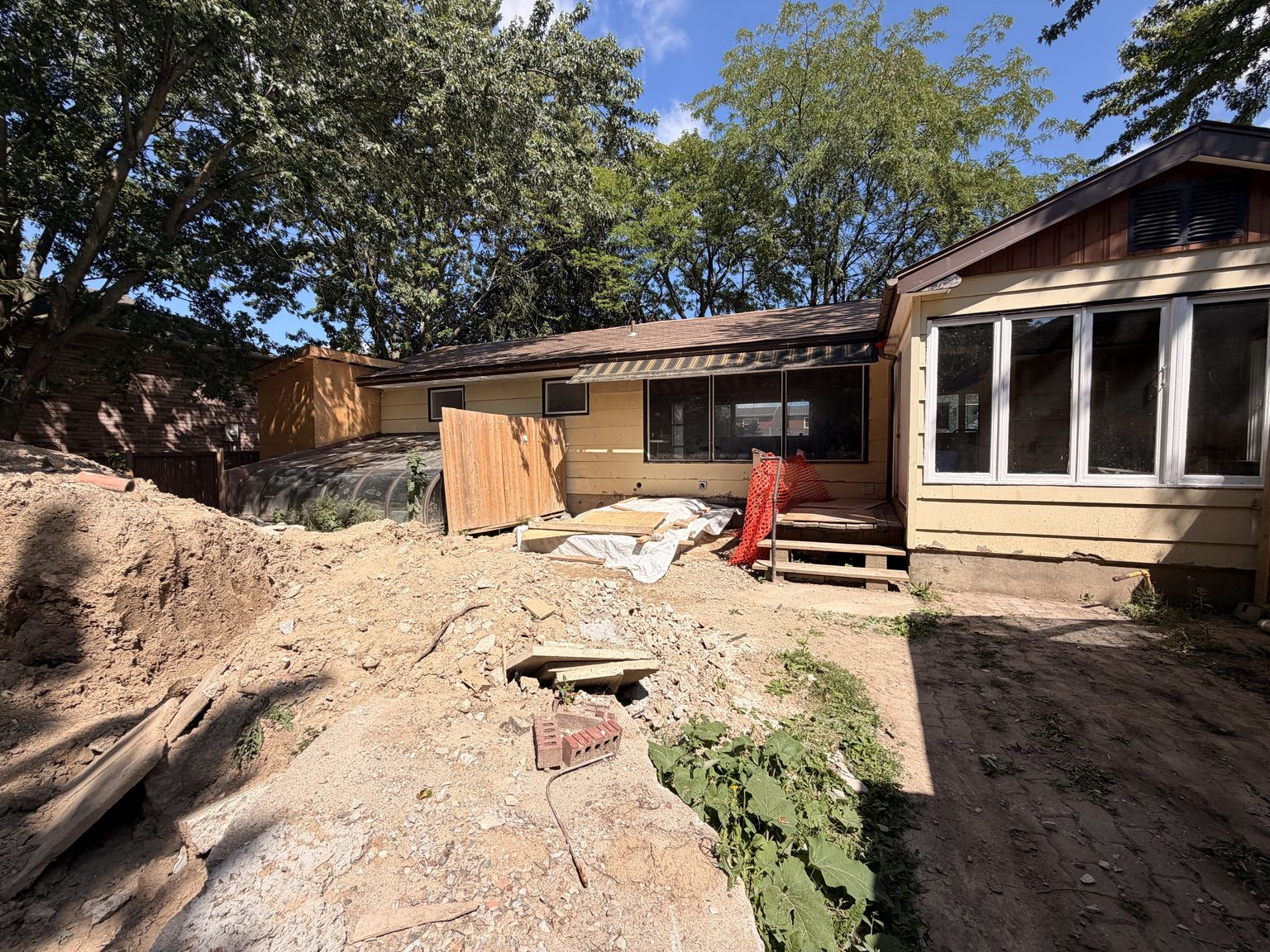 The back of 976 Stone Church Rd E under a blue sky, mature trees overhead, the back steps in shade and excavated ground across the foreground.