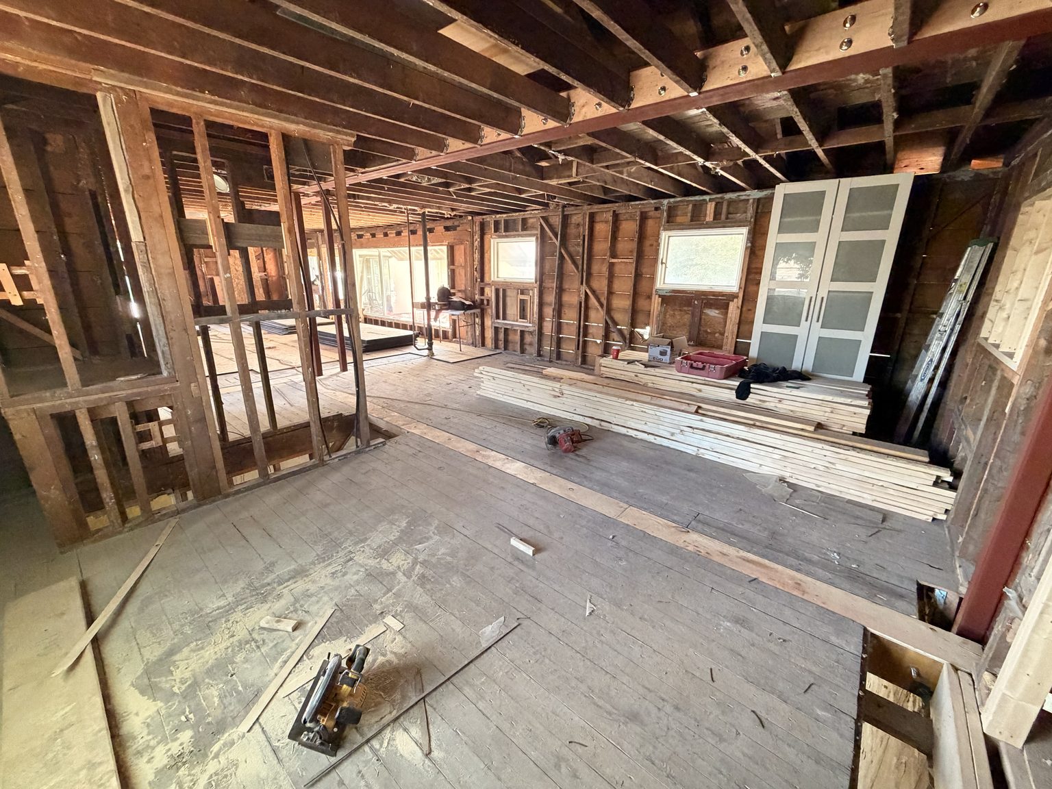 A program room stripped back to the timber frame, daylight coming through three windows, with a long stack of new framing lumber along the far wall.